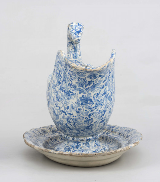 Sosjerka łódkowata z szerokim lekko wywiniętym wylewem, uchwyt wysoki w kształcie plastycznie modelowanej ptasiej głowy; zespolona z podstawką w formie owalnego talerzyka  z profilowanym kołnierzem. Powierzchnia pokryta białą glazurą , na niej niebieski nadruk z wzorem tzw. tapetowym złożonym ze stylizowanych motywów roślinnych. 
 