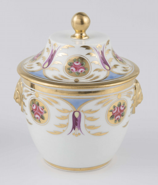 S/CS/1388/2/ML - Cukiernica półowoidalna z uchwytami w formie półplastycznych lwich głów i pokrywką w kształcie ściętego stożka o wklęsłych bokach z kulistym uchwytem. Dekoracja malowana wielobarwnie i złocona: brzusiec i boki pokrywki obiega szeroki fryz złożony ze stylizowanych ornamentów roślinnych (gałązek, kłosów zbóż i liści akantu)  połączonych z owalnymi medalionami, w których są trzy różowe różyczki z zielonymi listkami na złotym tle.Uchwyty malowane złotem,  złotym konturem obwiedzione wszystkie krawędzie.
 
