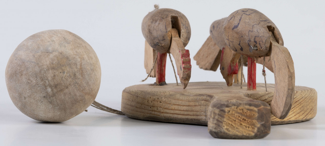 E/17030/ML - Zabawka drewniana przedstawiająca trzy kurki przymocowane do owalnej deseczki z uchwytem. Kurki mają ruchome głowy i ogony, które poruszają się za pomocą sznurków przeciągniętych przez deseczkę i przywiązanych do drewnianej kuli. Deseczka wykonana z drewna sosnowego, kurki z drewna lipowego. Głowy i nogi kurek pomalowane są czerwoną farbą, tułowia zdobi ornament geometryczny w postaci zygzakowatych linii wykonanych brązową farbą.