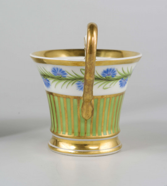 Filiżanka koniczna na pierścieniowatej stopie, ucho w formie pastorału z wolutą wyciągniętą ponad krawędź wylewu. Dekoracja malowana i złocona pokrywa całą powierzchnię: wokół wylewu fryz w postaci girlandy błękitnych chabrów z zielonymi listkami na białym tle, poniżej wzór pionowych pasków naprzemian złotych i zielonych; stopa, wylew (obustronnie) i ucho podkreślone złotymi paskami. 