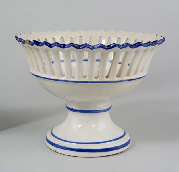Patera w formie kielicha na okrągłej nóżce, czara w górnej części ażurowa z pionowych pasków zamkniętych ząbkowanym brzegiem. Pokryta białym szkliwem, profile i krawędzie podkreślone paskami w kolorze błękitu kobaltowego.
 