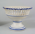 Patera w formie kielicha na okrągłej nóżce, czara w górnej części ażurowa z pionowych pasków zamkniętych ząbkowanym brzegiem. Pokryta białym szkliwem, profile i krawędzie podkreślone paskami w kolorze błękitu kobaltowego.
 