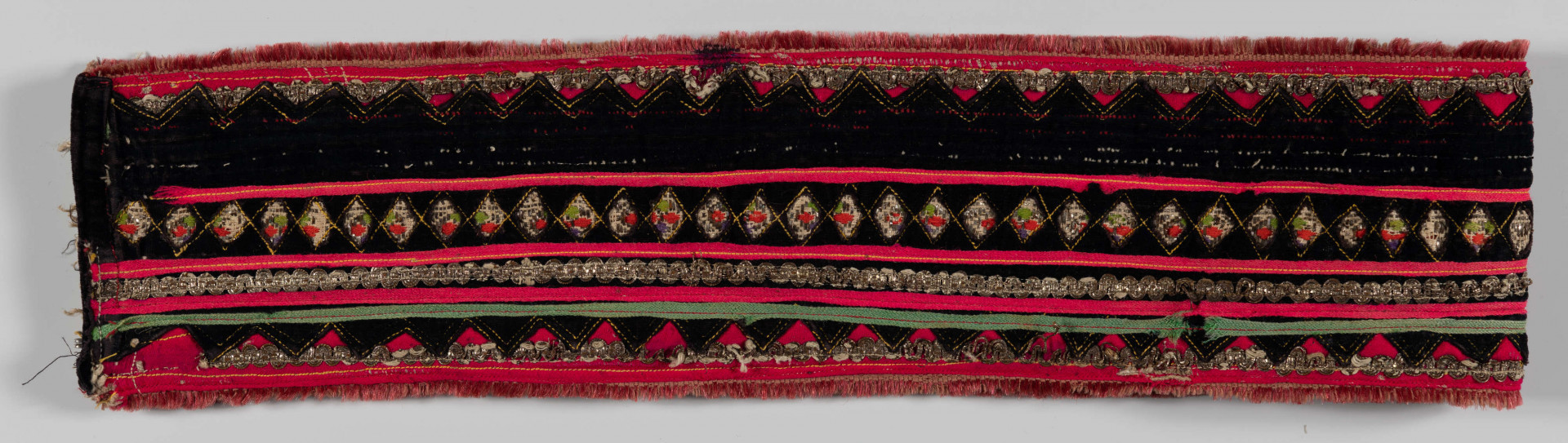 E/10999/ML - Pas męski szyty maszynowo z grubego lnianego płótna. Obszyty szczoteczką w kolorze czerwonym oraz cienką wełną w kolorze amarantowym i taśmą srebrzystą. Na środek naszyty jest czarny aksamit wycięty „w okienka” – ażurowy szlak z rombów. Pod okienka podłożone taśmy biało-czerwono-zielone ze srebrna nitką oraz amarantowe i zielone. Zapinany na 3 ciemne guziki i pętelki ręcznie wykonane z nici. 