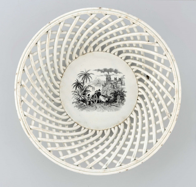 Koszyczek okrągły, szkliwiony, biały, dno gładkie, kołnierz ażurowy z okrągłych ukośnie wygiętych wałków ułożonych  w dwóch warstwach przciwlegle, zebranych potrójnym wałkiem tworzącym krawędź .W lustrze dekoracja w technice stalorytu w  kolorze czarnym: scenka rodzajowa w pejzażu o cechach południowych (palmy) z architekturą, w centrum dwukołowy wóz ciągnięty przez woły i postacie ludzi.
 
