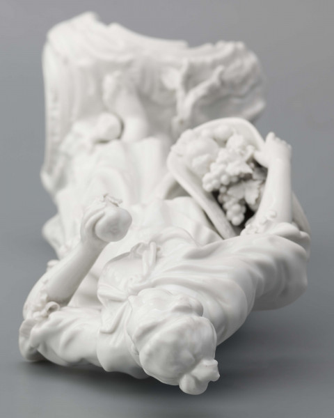 S/CS/1476/ML - Figurka z białej szkliwionej porcelany przedstawiajaca  kobietę stojacąna niskim trójdzielnym cokole. Kobieta ma długie włosy upięte w koczek, ubrana jest w fałdzistą spódnicę przewiązaną poniżej bioder, gorset z baskinką i głębokim dekoltem, bluzkę z bufiastmi rękawami do łokci, na ramionach zawiazaną chustkę, ma bose stopy. Prawą ręką trzyma płaski kosz z owocami oparty na biodrze, w lewej ugiętej ma jabłko. 
  