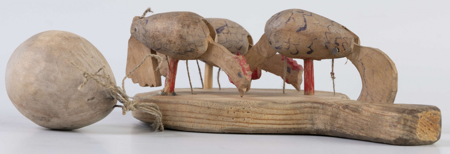 E/17030/ML - Zabawka drewniana przedstawiająca trzy kurki przymocowane do owalnej deseczki z uchwytem. Kurki mają ruchome głowy i ogony, które poruszają się za pomocą sznurków przeciągniętych przez deseczkę i przywiązanych do drewnianej kuli. Deseczka wykonana z drewna sosnowego, kurki z drewna lipowego. Głowy i nogi kurek pomalowane są czerwoną farbą, tułowia zdobi ornament geometryczny w postaci zygzakowatych linii wykonanych brązową farbą.