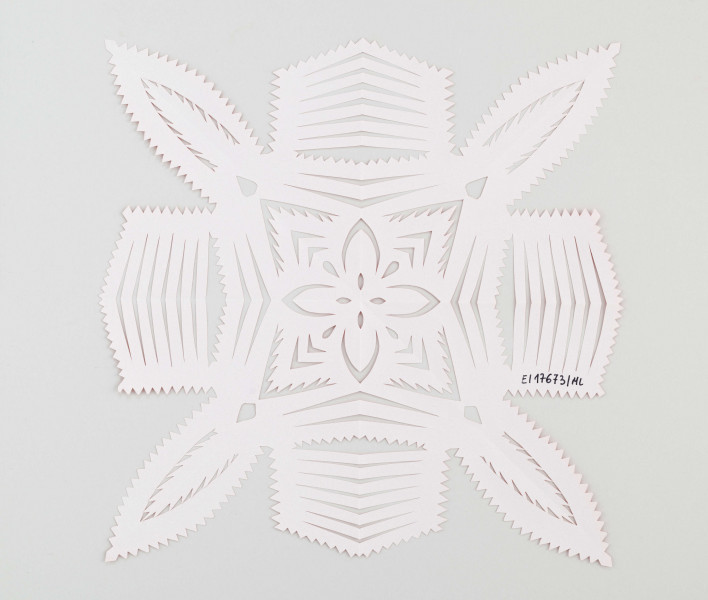 Wycinanka koloru czerwonego, przypominająca nałożone na siebie dwa krzyże; jeden o szerszych ramionach, drugi o ramionach płatkowych. Wokół wykończone ząbkami. W środku kompozycja z płatków i  strzałek, przypominająca kwiat. 