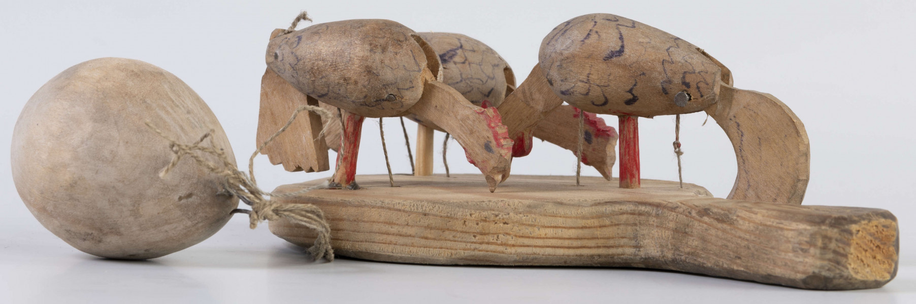 E/17030/ML - Zabawka drewniana przedstawiająca trzy kurki przymocowane do owalnej deseczki z uchwytem. Kurki mają ruchome głowy i ogony, które poruszają się za pomocą sznurków przeciągniętych przez deseczkę i przywiązanych do drewnianej kuli. Deseczka wykonana z drewna sosnowego, kurki z drewna lipowego. Głowy i nogi kurek pomalowane są czerwoną farbą, tułowia zdobi ornament geometryczny w postaci zygzakowatych linii wykonanych brązową farbą.