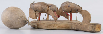 E/17030/ML - Zabawka drewniana przedstawiająca trzy kurki przymocowane do owalnej deseczki z uchwytem. Kurki mają ruchome głowy i ogony, które poruszają się za pomocą sznurków przeciągniętych przez deseczkę i przywiązanych do drewnianej kuli. Deseczka wykonana z drewna sosnowego, kurki z drewna lipowego. Głowy i nogi kurek pomalowane są czerwoną farbą, tułowia zdobi ornament geometryczny w postaci zygzakowatych linii wykonanych brązową farbą.