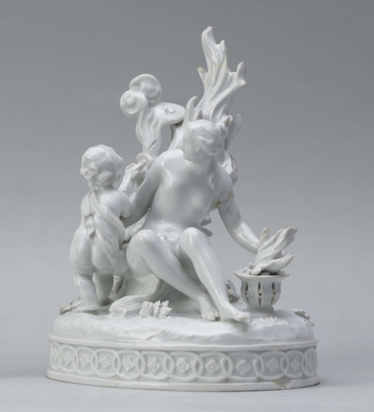 S/CS/1456/ML - Grupa figuralna z białej szkliwionej porcelany. Cokół owalny, wysklepiony z ornamentem opaskowym złożonym ze splecionych owali z rozetkami. Na nim siedzi naga kobieta (Venus) przed płonącym stosem o wysokich płomieniach i wyciągniętą w bok prawą ręką powstrzymuje nagie dziecko (Amorek) zapalające od ognia pochodnię, lewą ręką siega do ażurowego naczynia z ogniem. Za nią na powierzchni cokołu drobne roślinki.
