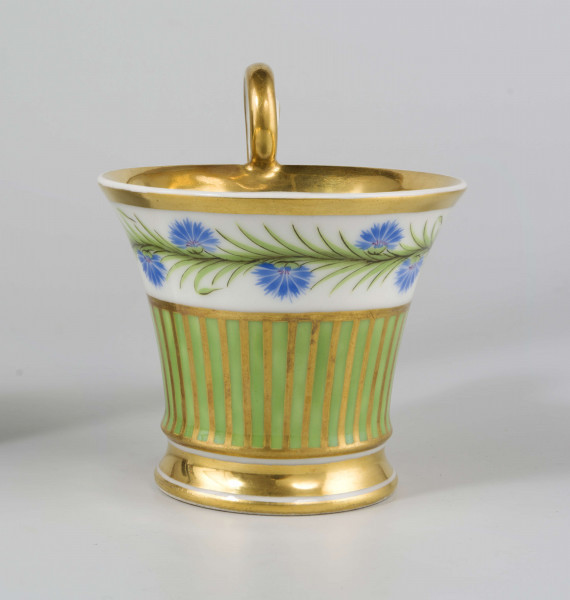 Filiżanka koniczna na pierścieniowatej stopie, ucho w formie pastorału z wolutą wyciągniętą ponad krawędź wylewu. Dekoracja malowana i złocona pokrywa całą powierzchnię: wokół wylewu fryz w postaci girlandy błękitnych chabrów z zielonymi listkami na białym tle, poniżej wzór pionowych pasków naprzemian złotych i zielonych; stopa, wylew (obustronnie) i ucho podkreślone złotymi paskami. 