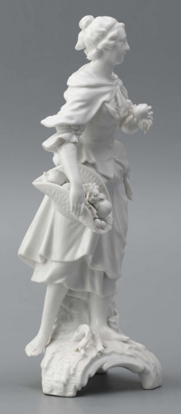 S/CS/1476/ML - Figurka z białej szkliwionej porcelany przedstawiajaca  kobietę stojacąna niskim trójdzielnym cokole. Kobieta ma długie włosy upięte w koczek, ubrana jest w fałdzistą spódnicę przewiązaną poniżej bioder, gorset z baskinką i głębokim dekoltem, bluzkę z bufiastmi rękawami do łokci, na ramionach zawiazaną chustkę, ma bose stopy. Prawą ręką trzyma płaski kosz z owocami oparty na biodrze, w lewej ugiętej ma jabłko. 
  