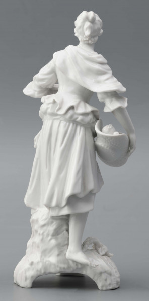 S/CS/1476/ML - Figurka z białej szkliwionej porcelany przedstawiajaca  kobietę stojacąna niskim trójdzielnym cokole. Kobieta ma długie włosy upięte w koczek, ubrana jest w fałdzistą spódnicę przewiązaną poniżej bioder, gorset z baskinką i głębokim dekoltem, bluzkę z bufiastmi rękawami do łokci, na ramionach zawiazaną chustkę, ma bose stopy. Prawą ręką trzyma płaski kosz z owocami oparty na biodrze, w lewej ugiętej ma jabłko. 
  