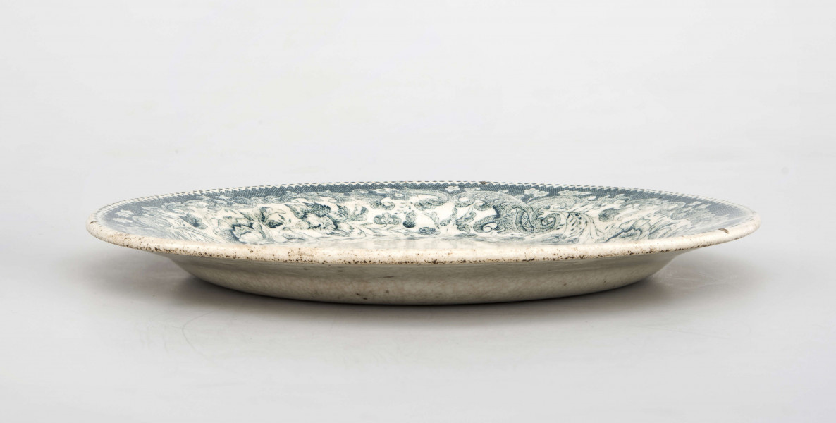 Talerz deserowy, okrągły, gładki, pokryty szkliwem białym, na nim dekoracja  drukowana w kolorze zieleni chromowej: w lustrze scena rodzajowa - kobieta z bawiącym sie dzieckiem; na kołnierzu szeroka bordiura z motywem ząbkowanej koronki, ornamentów wolutowych i kwiatów. 
 