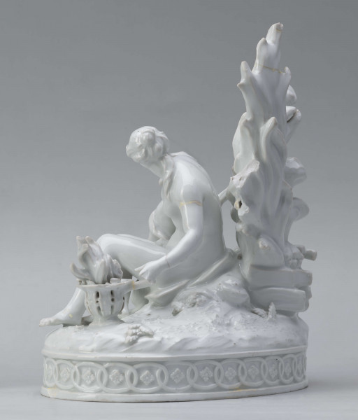S/CS/1456/ML - Grupa figuralna z białej szkliwionej porcelany. Cokół owalny, wysklepiony z ornamentem opaskowym złożonym ze splecionych owali z rozetkami. Na nim siedzi naga kobieta (Venus) przed płonącym stosem o wysokich płomieniach i wyciągniętą w bok prawą ręką powstrzymuje nagie dziecko (Amorek) zapalające od ognia pochodnię, lewą ręką siega do ażurowego naczynia z ogniem. Za nią na powierzchni cokołu drobne roślinki.
