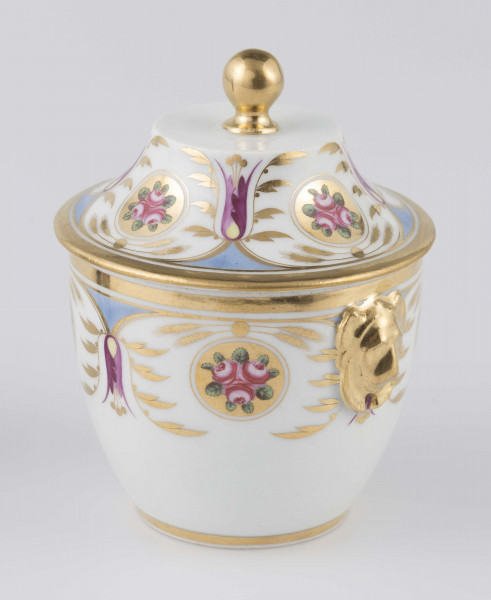 S/CS/1388/2/ML - Cukiernica półowoidalna z uchwytami w formie półplastycznych lwich głów i pokrywką w kształcie ściętego stożka o wklęsłych bokach z kulistym uchwytem. Dekoracja malowana wielobarwnie i złocona: brzusiec i boki pokrywki obiega szeroki fryz złożony ze stylizowanych ornamentów roślinnych (gałązek, kłosów zbóż i liści akantu)  połączonych z owalnymi medalionami, w których są trzy różowe różyczki z zielonymi listkami na złotym tle.Uchwyty malowane złotem,  złotym konturem obwiedzione wszystkie krawędzie.
 