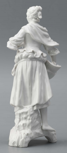 S/CS/1476/ML - Figurka z białej szkliwionej porcelany przedstawiajaca  kobietę stojacąna niskim trójdzielnym cokole. Kobieta ma długie włosy upięte w koczek, ubrana jest w fałdzistą spódnicę przewiązaną poniżej bioder, gorset z baskinką i głębokim dekoltem, bluzkę z bufiastmi rękawami do łokci, na ramionach zawiazaną chustkę, ma bose stopy. Prawą ręką trzyma płaski kosz z owocami oparty na biodrze, w lewej ugiętej ma jabłko. 
  