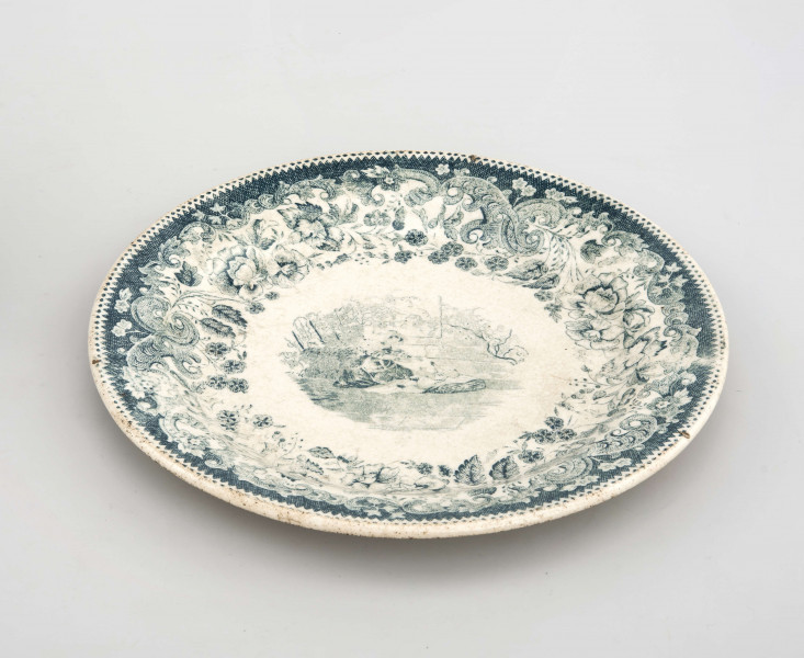 Talerz deserowy, okrągły, gładki, pokryty szkliwem białym, na nim dekoracja  drukowana w kolorze zieleni chromowej: w lustrze scena rodzajowa - kobieta z bawiącym sie dzieckiem; na kołnierzu szeroka bordiura z motywem ząbkowanej koronki, ornamentów wolutowych i kwiatów. 
 