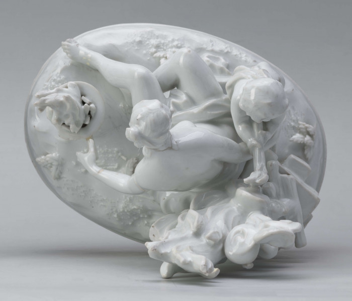 S/CS/1456/ML - Grupa figuralna z białej szkliwionej porcelany. Cokół owalny, wysklepiony z ornamentem opaskowym złożonym ze splecionych owali z rozetkami. Na nim siedzi naga kobieta (Venus) przed płonącym stosem o wysokich płomieniach i wyciągniętą w bok prawą ręką powstrzymuje nagie dziecko (Amorek) zapalające od ognia pochodnię, lewą ręką siega do ażurowego naczynia z ogniem. Za nią na powierzchni cokołu drobne roślinki.
