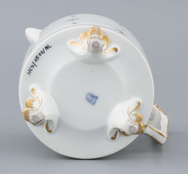 S/CS/1511/ML - Dzbanek cylindryczny, na trzech nóżkach w kształcie lwich łap, szyja w formie zwężonego pierścienia, wylew z dziobkiem, ucho dołem prostokątnie załamane, górą wygięte, rozdwojone, z reliefowymi liśćmi w osadzeniach. Przykrywa w formie wąskiego, konicznego pierścienia, z uchwytem w kształcie szyszki. Dekoracja malowana: regularny rzut drownych chabrów w kolorach błękitu i zieleni. Krawędzie,  ucho, dziobek, nóżki i uchwyt pokrywki konturowane złotem.
