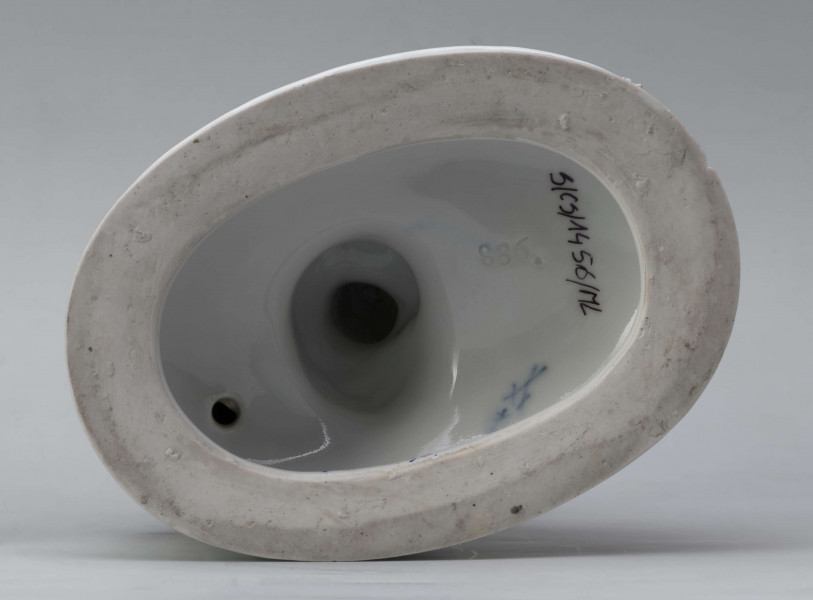 S/CS/1456/ML - Grupa figuralna z białej szkliwionej porcelany. Cokół owalny, wysklepiony z ornamentem opaskowym złożonym ze splecionych owali z rozetkami. Na nim siedzi naga kobieta (Venus) przed płonącym stosem o wysokich płomieniach i wyciągniętą w bok prawą ręką powstrzymuje nagie dziecko (Amorek) zapalające od ognia pochodnię, lewą ręką siega do ażurowego naczynia z ogniem. Za nią na powierzchni cokołu drobne roślinki.
