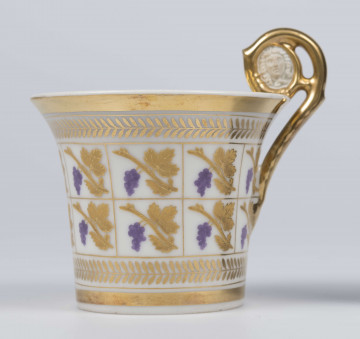 S/CS/136/1/ML - Filiżanka koniczna, ucho w formie pastorału z wolutą wyciągniętą ponad krawędź wylewu zdobioną reliefowym  medalionem z głową meduzy. Dekoracja malowana złotem z dodatkiem fioletowej purpury pokrywa całą powierzchnię: przez środek biegnie dwurzędowazłota  kratownica wypełniona motywem winogrona na złotej gałązce, obramowana wicią złotych listków, dolna i górna krawędź obwiedzione złotymi paskami, złocone ucho i wnętrze wylewu. 