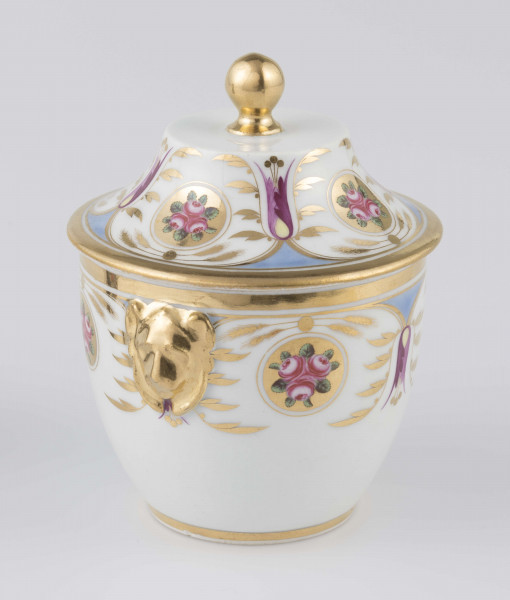 S/CS/1388/2/ML - Cukiernica półowoidalna z uchwytami w formie półplastycznych lwich głów i pokrywką w kształcie ściętego stożka o wklęsłych bokach z kulistym uchwytem. Dekoracja malowana wielobarwnie i złocona: brzusiec i boki pokrywki obiega szeroki fryz złożony ze stylizowanych ornamentów roślinnych (gałązek, kłosów zbóż i liści akantu)  połączonych z owalnymi medalionami, w których są trzy różowe różyczki z zielonymi listkami na złotym tle.Uchwyty malowane złotem,  złotym konturem obwiedzione wszystkie krawędzie.
 