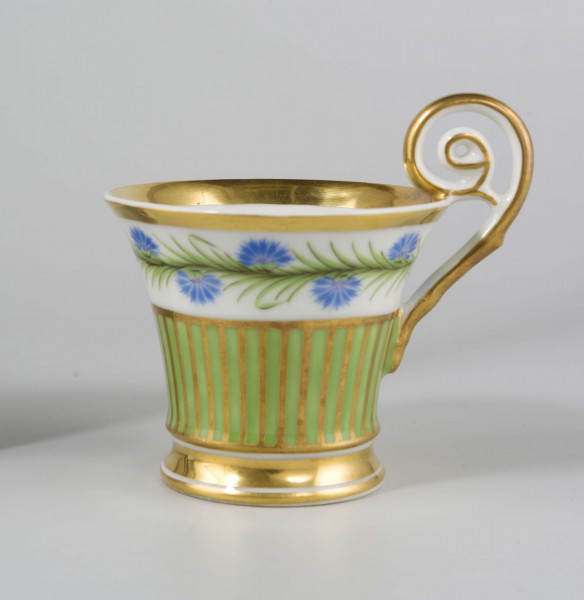 Filiżanka koniczna na pierścieniowatej stopie, ucho w formie pastorału z wolutą wyciągniętą ponad krawędź wylewu. Dekoracja malowana i złocona pokrywa całą powierzchnię: wokół wylewu fryz w postaci girlandy błękitnych chabrów z zielonymi listkami na białym tle, poniżej wzór pionowych pasków naprzemian złotych i zielonych; stopa, wylew (obustronnie) i ucho podkreślone złotymi paskami. 