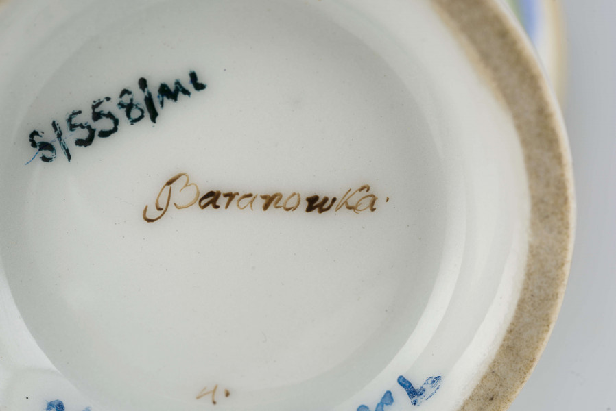 Filiżanka koniczna na pierścieniowatej stopie, ucho w formie pastorału z wolutą wyciągniętą ponad krawędź wylewu. Dekoracja malowana i złocona pokrywa całą powierzchnię: wokół wylewu fryz w postaci girlandy błękitnych chabrów z zielonymi listkami na białym tle, poniżej wzór pionowych pasków naprzemian złotych i zielonych; stopa, wylew (obustronnie) i ucho podkreślone złotymi paskami. 