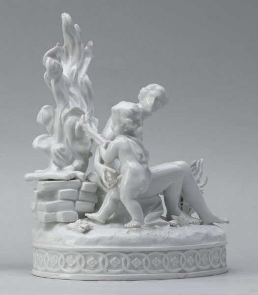 S/CS/1456/ML - Grupa figuralna z białej szkliwionej porcelany. Cokół owalny, wysklepiony z ornamentem opaskowym złożonym ze splecionych owali z rozetkami. Na nim siedzi naga kobieta (Venus) przed płonącym stosem o wysokich płomieniach i wyciągniętą w bok prawą ręką powstrzymuje nagie dziecko (Amorek) zapalające od ognia pochodnię, lewą ręką siega do ażurowego naczynia z ogniem. Za nią na powierzchni cokołu drobne roślinki.
