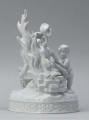 S/CS/1456/ML - Grupa figuralna z białej szkliwionej porcelany. Cokół owalny, wysklepiony z ornamentem opaskowym złożonym ze splecionych owali z rozetkami. Na nim siedzi naga kobieta (Venus) przed płonącym stosem o wysokich płomieniach i wyciągniętą w bok prawą ręką powstrzymuje nagie dziecko (Amorek) zapalające od ognia pochodnię, lewą ręką siega do ażurowego naczynia z ogniem. Za nią na powierzchni cokołu drobne roślinki.
