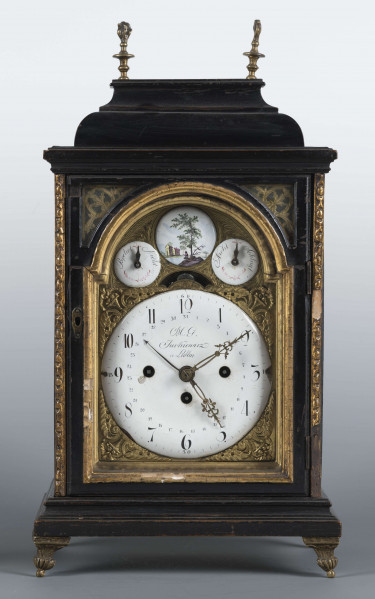 S/Mt/531/ML - Obudowa szafkowa, czworoboczna ze schodkowym zwieńczeniem, pokryta czarną politurą, oszklona z czterech stron, na czterech nóżkach z mosiądzu; w górnych narożnikach mosiężne sterczyny w formie płonących wazonów. Mechanizm chodu i bicia o napędzie sprężynowym, z wahadłem i dwoma dzwonkami; bicie godzin i kwadransów, budzik i repetier. Tarcza biało emaliowana z czarnymi cyframi arabskimi godzin i minut, w niej trzy otwory na osie sprężyn; trzy wskazówki, godzinowa i minutowa - ozdobne, sekundowa prosta. W górnej części tarczy sygnatura pisana ozdobną kursywą: M.G. / Jurkiewicz /  w Lublinie. Powyżej tarczy wycięcie z widocznym wahadełkiem kontrolnym i trzy mniejsze emaliowane tarcze: środkowa z miniaturą pejzażową, boczne z wyłączeniem chodu i bicia.