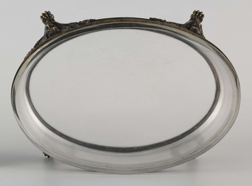 S/CS/1063/ML - Naczynie ze szkła bezbarwnego, grubościennego, owalne, z płaskim dnem, w oprawie metalowej złożonej z dwóch obręczy połączonych plastycznymi figurkami gryfów na nóżkach. Powierzchnia obręczy zdobiona plastycznym fryzem antykizujacych palmet. Oprawa metalowa patynowana. 
 