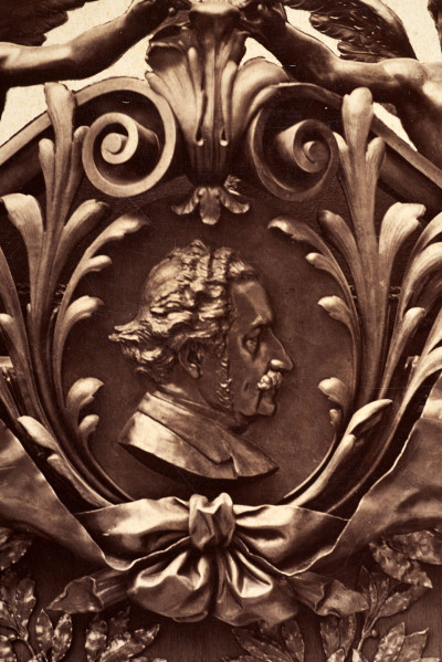 Pośrodku prostokątnego pola o zaokrąglonych narożnikach większa fotografia przyozdobiona wstęgą i gałązkami wawrzynu. Dookoła szesnaście symetrycznie rozmieszczonych mniejszych fotografii. W prawym górnym rogu dr Wacław Lasocki, naczelny lekarz kolei nadwiślańskiej w latach 1875 - 78. W polu ramy w górnej części medalion z płaskorzeźbionym popiersiem z profilu. Przedstawia Leopolda Kronenberga - ojca. Po bokach dwia półleżące aniołki trzymają wieniec laurowy. Po lewej stronie ramy postać robotnika Poleszuka, po prawej pracownika warsztatów kolejowych. W dolnej części front lokomotywy i złoty żeton uprawniający do bezpłatnej jazdy pociągiem I klasy. Fotografia naklejona na tekturę, w dolnej części u dołu sucha pieczęć zakładu fotograficznego.