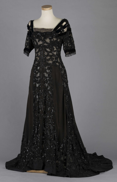 S/T/243/ML - Suknia wizytowa, czarna, uszyta z atłasu jedwabnego, jedwabnego tiulu i aksamitu, haftowana cekinami. Krótki rękaw z jedwabnego czarnego aksamitu, dekolt prostokatny, dość głęboki, wykończony wstawką haftowaną i aksamitem. Kolory są kontrastowe. Spodnia warstwa sukni, z białej tafty i muślinu, widoczna w partiach sukni szytych z czarnego tiulu, dodaje całości elegancji. Brzegi dekoracyjnej wici z tafty zaakcentowane naszytymi cekinami. Suknia do ziemi z tyłu krótki tren. Brak halki. 