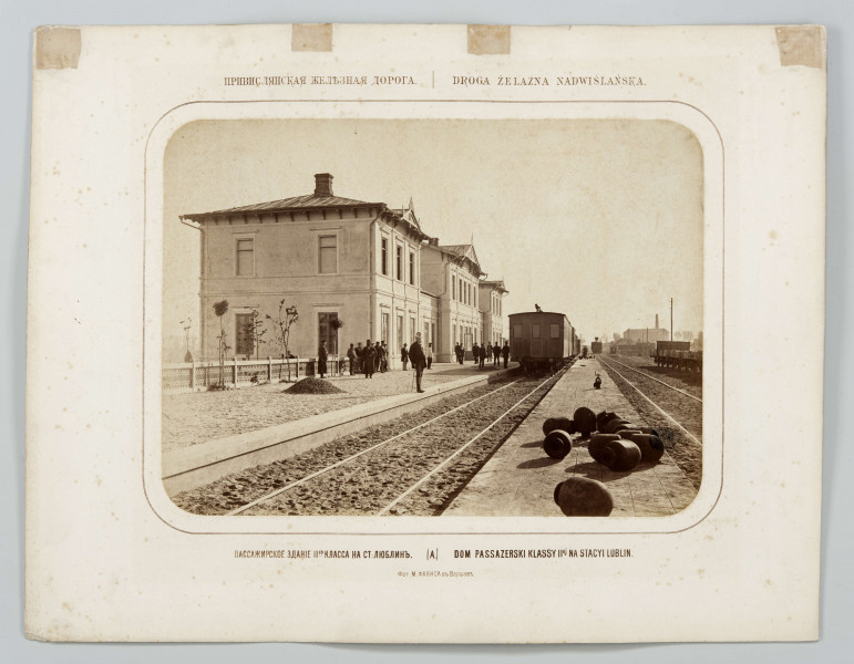 Fotografia sepiowana dworca kolejowego w Lublinie - widok budynku pasażerskiego od strony torów. Po lewej stronie peron i budynek dworca. Pośrodku na pierwszym planie ułożone pomiędzy torami owalne dużych gabarytów przedmioty. W oddali wagony na torach. Po prawej fragment wagonu i budynki w tle. Główny budynek stacji murowany, piętrowy. Dach kryty, szczyty zwieńczone ozdobną snycerką. Widoczne przęsła ogrodzenia oddzielającego plac przez budynkiem od peronu. Obok kilka małych drzew. Na peronie kilkunastu mężczyzn w strojach z epoki.
Fotografia o zaokrąglonych rogach, obrysowana dookoła cienką ramką, naklejona na tekturę. Nad i pod fotografią dwujęzyczne rosyjsko-polskie napisy informacyjne. W dolnej części karty w języku rosyjskim informacja o autorze zdjęcia.
