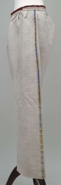 E/12943/ML - Spodnie z lnianego materiału tkanego splotem prostym. Szyte ręcznie i na maszynie. Nogawki szerokie ujęte w pasek szer. 3 cm, wzmocniony i ozdobiony paskiem płótna z ornamentem tkackim „pereborami”. Poniżej paska wszyte 2 trójkątne kliny oraz podobny w rozporku, zapinanym na 2 guziki. Na szwach bocznych nogawek naszyte kolorowe, fabryczne tasiemki, szer. 1,5 cm – żółta i niebieska.