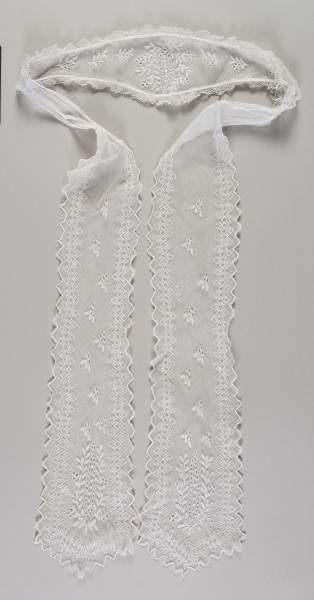 E/16857/ML - Czepek tiulowy biały, składający się z łówki w kształcie diademu oraz 2 rozszerzających się ku końcowi wiązadeł. Na każdym elemencie fabryczne hafty z białej nici. Krawędzie główki obszyte podwójną falbanką z ręcznie wykonanej koronki. Na całej długości głowi hafty z motywem wielopłatkowych kwiatów z łodygami i liśćmi. Pośrodku kwiatostan z pojedynczych pączków otoczony gałązką z liśćmi i pączkami. Krawędzie wiązadeł wycięte w ząbki i wykończone ściegiem obrębiającym. Na powierzchni haft z motywem pojedynczych gałązek z pączkami i liśćmi, w dolnej części – kwiatostan podobny do tego na główce. Wiązadła ozdobione dodatkowo bordiurą  z ząbków.