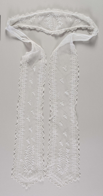 E/16857/ML - Czepek tiulowy biały, składający się z łówki w kształcie diademu oraz 2 rozszerzających się ku końcowi wiązadeł. Na każdym elemencie fabryczne hafty z białej nici. Krawędzie główki obszyte podwójną falbanką z ręcznie wykonanej koronki. Na całej długości głowi hafty z motywem wielopłatkowych kwiatów z łodygami i liśćmi. Pośrodku kwiatostan z pojedynczych pączków otoczony gałązką z liśćmi i pączkami. Krawędzie wiązadeł wycięte w ząbki i wykończone ściegiem obrębiającym. Na powierzchni haft z motywem pojedynczych gałązek z pączkami i liśćmi, w dolnej części – kwiatostan podobny do tego na główce. Wiązadła ozdobione dodatkowo bordiurą  z ząbków.