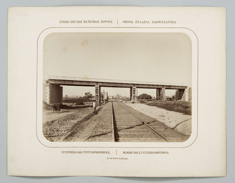 Przy nasypie po lewej stronie torów stoi mężczyzna, na lewo wóz. Po drugiej stronie pod wiaduktem widoczna grupa mężczyzn. W tle zabudowania kolejowe, drzewa i fragment miasta. Fotografia o zaokrąglonych rogach, obrysowana dookoła cienką ramką, naklejona na tekturę. Powyżej i pod fotografią napisy po rosyjsku - z lewej i po polsku - z prawej strony. W dolnej części informacja o autorze zdjęcia w języku rosyjskim.