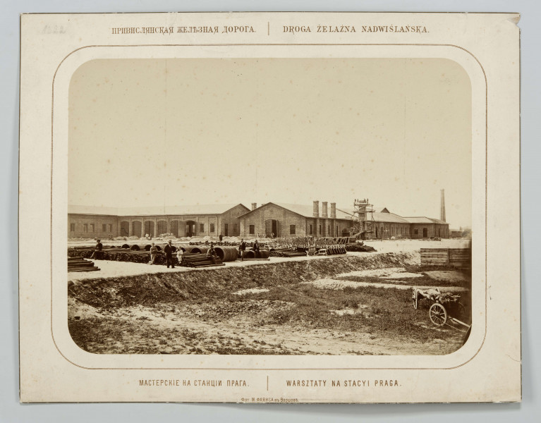 Budynki warsztatów kolejowych, parterowe, nakryte dachem dwuspadowym. Na placu przed budynkami złożone materiały budowlane: rury różnego przekroju, taczki, pomiędzy nimi kilku mężczyzn. W prawym dolnym rogu wóz. Przy budynku rusztowanie, w oddali komin. Fotografia o zaokrąglonych rogach, obrysowana dookoła cienką ramką, naklejona na tekturę, na brzegu tektury ramka. Powyżej i pod fotografią napisy po rosyjsku - z lewej i po polsku - z prawej strony. W dolnej części informacja o autorze zdjęcia po rosyjsku.