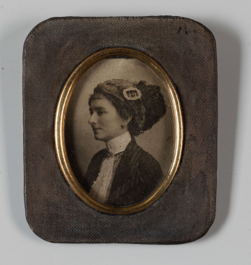 MPol/173/ML - Fotografia Idalii Polówny, wnuczki Wincentego Pola, córki Marka Stanisława Pola, syna poety. Fotografia przedstawia popiersie młodej kobiety, jej lewy półprofil. Kobieta ubrana w fantazyjne nakrycie głowy i ciemny płaszcz z jasnym żabotem, zapięty na białej stójce. Pod żabotem widać bluzkę w kratkę. Włosy ciemne, upięte. Fotografia umieszczona w mosiężnej oprawie, z owalnym miejscem na zdjęcie. Ramka prostokątna, z zaokrąglonymi narożnikami.