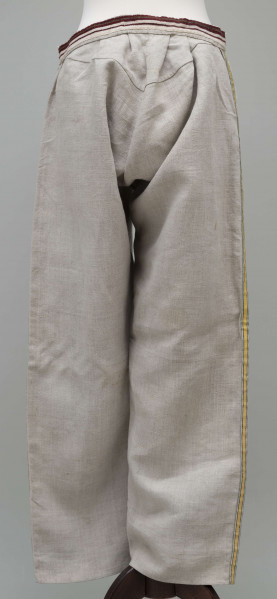 E/12943/ML - Spodnie z lnianego materiału tkanego splotem prostym. Szyte ręcznie i na maszynie. Nogawki szerokie ujęte w pasek szer. 3 cm, wzmocniony i ozdobiony paskiem płótna z ornamentem tkackim „pereborami”. Poniżej paska wszyte 2 trójkątne kliny oraz podobny w rozporku, zapinanym na 2 guziki. Na szwach bocznych nogawek naszyte kolorowe, fabryczne tasiemki, szer. 1,5 cm – żółta i niebieska.