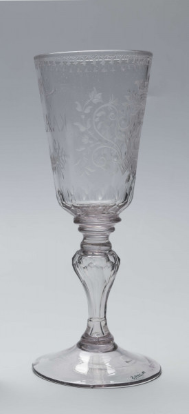 S/CS/244/ML - Stopa okrągła, lekko uniesiona, trzon tralkowy, z pęcherzykiem powietrza, fasetowany, od czaszy oddzielony pierścieniem i dwoma talerzykami, czasza stozkowata, u nasady zaokrąglona, szlifowana w dwa pionowe rzędy owalnych faset. Dekoracja rytowana: na czaszy dwustronna - z jednej strony owalna tarcza z parą gołąbków połączonych dziobkami stojących na serduszkach w rozbudowanym kartuszu z uszakami, z którego wyrastaja gałązki; po drugiej stronie litery 