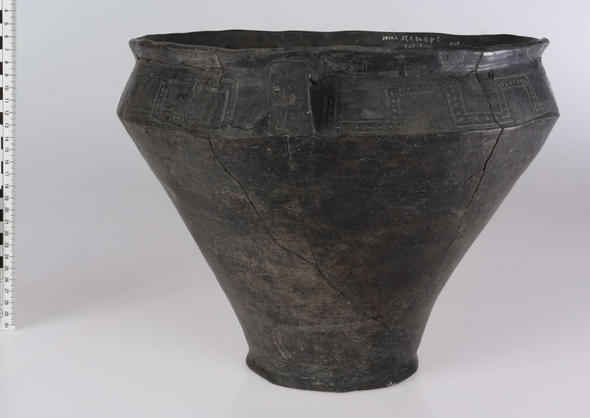 765/A/ML/1 - Waza wyraźnie profilowana, z prostym, odgietym na zewnatrz brzegiem, ostro zaznaczonym załomem brzuśca i wyodrębnionym dnem. Ponad załomem brzuśca występuje ornament w postaci: jodełek, swastyk, tworzonych przez dwie linie ryte i rząd dołków pomiędzy nimi oraz ornament blankowy. Naczynie ma trzy taśmowate ucha kolankowato zgięte. 
