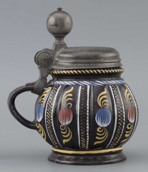 S/CS/595/ML - Kufel  kulisty, o niskiej konicznej stopie i niskiej szyi z pierścieniem, ucho taśmowe w kształcie „C”, pokrywka cynowa. Korpus pionowo kanelowany, podzielony na pasy na przemian gładkie i rytowane w ukośną kratkę. Pokryty szkliwem w kolorze ciemnego kobaltu, zdobiony malowanymi ornamentami podkreślającymi podziały powierzchni, w kolorach: czerwień, żółcień, błękit, biel. Pokrywka płaska, z pierścieniami i uchwytem kulkowym, na zawiasie umocowanym do ucha. Na wierzchu pokrywki grawerunek przedstawiający zawiły ornament – być może monogram spleciony z liter JGL – pod koroną, w uproszczonym wieńcu, z datą 1716.  