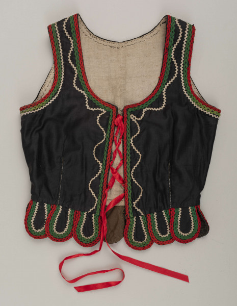 E/16876/ML - Gorset zszyty z 6 kawałków czarnej satyny, podszyty lnianą podszewką. Z przodu rozcięcie usztywnione fiszbinami oraz haftkami do przewlekania wstążeczki. Od pasa dookoła opadają klapki „taszki”. Przód wzdłuż rozcięcie wycięcie wokół szyi, brzegi pach i taszki ozdobione czerwoną, zieloną i białą wełnianą krepinką. Plecy zdobione ornamentem wykonanym ściegiem krzyżykowym. Szyty ręcznie.