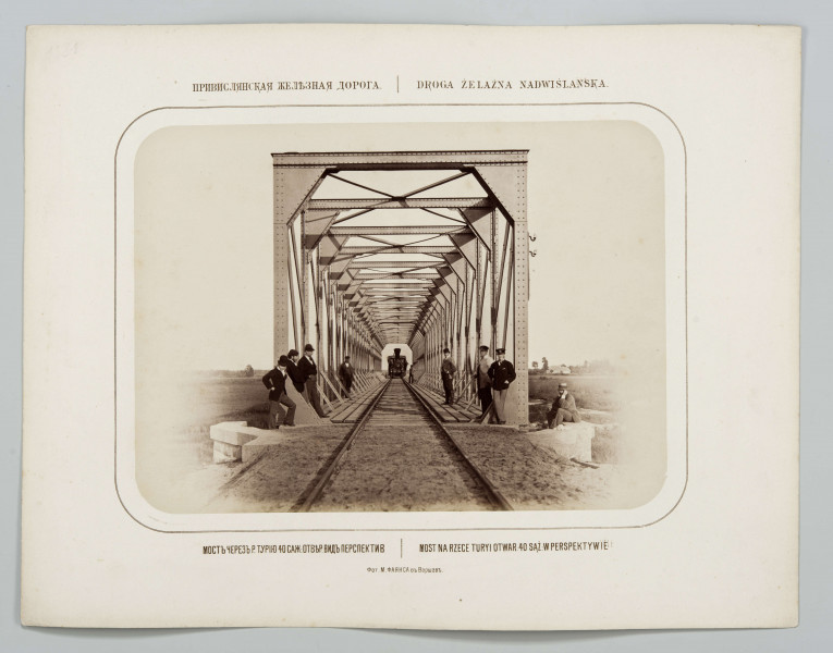 Widok wlotu mostu w perspektywie, przy metalowych słupach stoją mężczyźni, jeden siedzi na murku. Na drugim końcu mostu lokomotywa. W tle pejzaż wiejski z domem po prawej. Fotografia o zaokrąglonych rogach, obrysowana dookoła cienką ramką, naklejona na tekturę. Powyżej i pod fotografią napisy po rosyjsku - z lewej i po polsku- z prawej strony. W dolnej części informacja o autorze zdjęcia po rosyjsku.