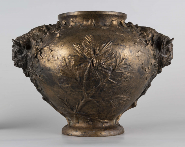 S/Mt/427/ML - Waza w kształcie spłaszczonej kuli, z niską, kolistą stopą i krótką szyją o lekko wywiniętym brzegu. Powierzchnia zdobiona reliefem: po bokach dwa uchwyty w formie głów żubrów, wokół nich gałązki dębu, pośrodku korpusu gałązki sosny z szyszkami wyrastające ze stopy. U góry na brzuścu rytowany napis majuskułą 