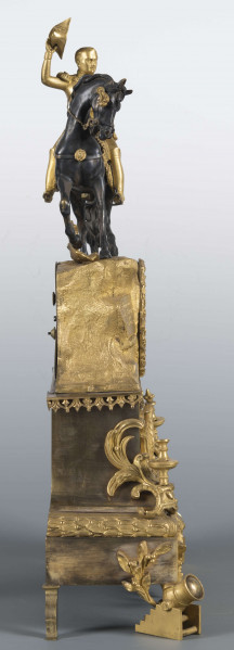 S/Mt/410/ML - Zegar kominkowy z brązu złoconego w stylu empire. Obudowa trzystopniowa, zwężająca się ku górze, na czterech nóżkach w formie armat, złożona z prostokątnej podstawy, części środkowej o wklęsłej linii boków i górnej imitującej skalisty szczyt; podziały podkreślone pasami ornamentu. Powierzchnię zdobią nakładki – w dolnej części panoplia i wieńce laurowe, w środkowej cesarski orzeł, w narożach umieszczono znicze. Pośrodku części górnej wieniec dębowy a w nim umieszczona okrągła tarcza zegarowa pokryta białą emalią, z czarnymi cyframi rzymskimi godzin i oznaczeniami minut. W tarczy dwie ażurowe wskazówki i dwa otwory na osie sprężyn, po bokach tarczy wyryte napisy: 